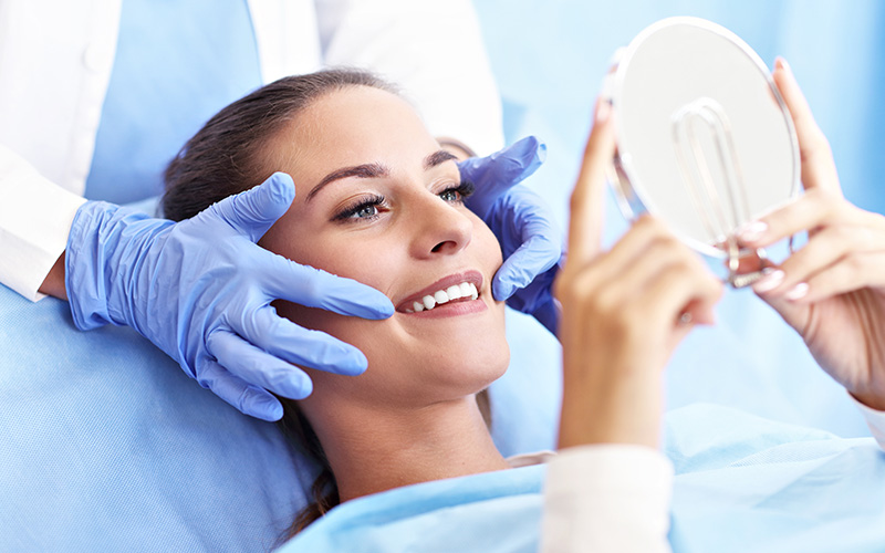 A person lying on a dental chair with a smile, receiving dental care from a professional wearing blue gloves and holding a mirror.